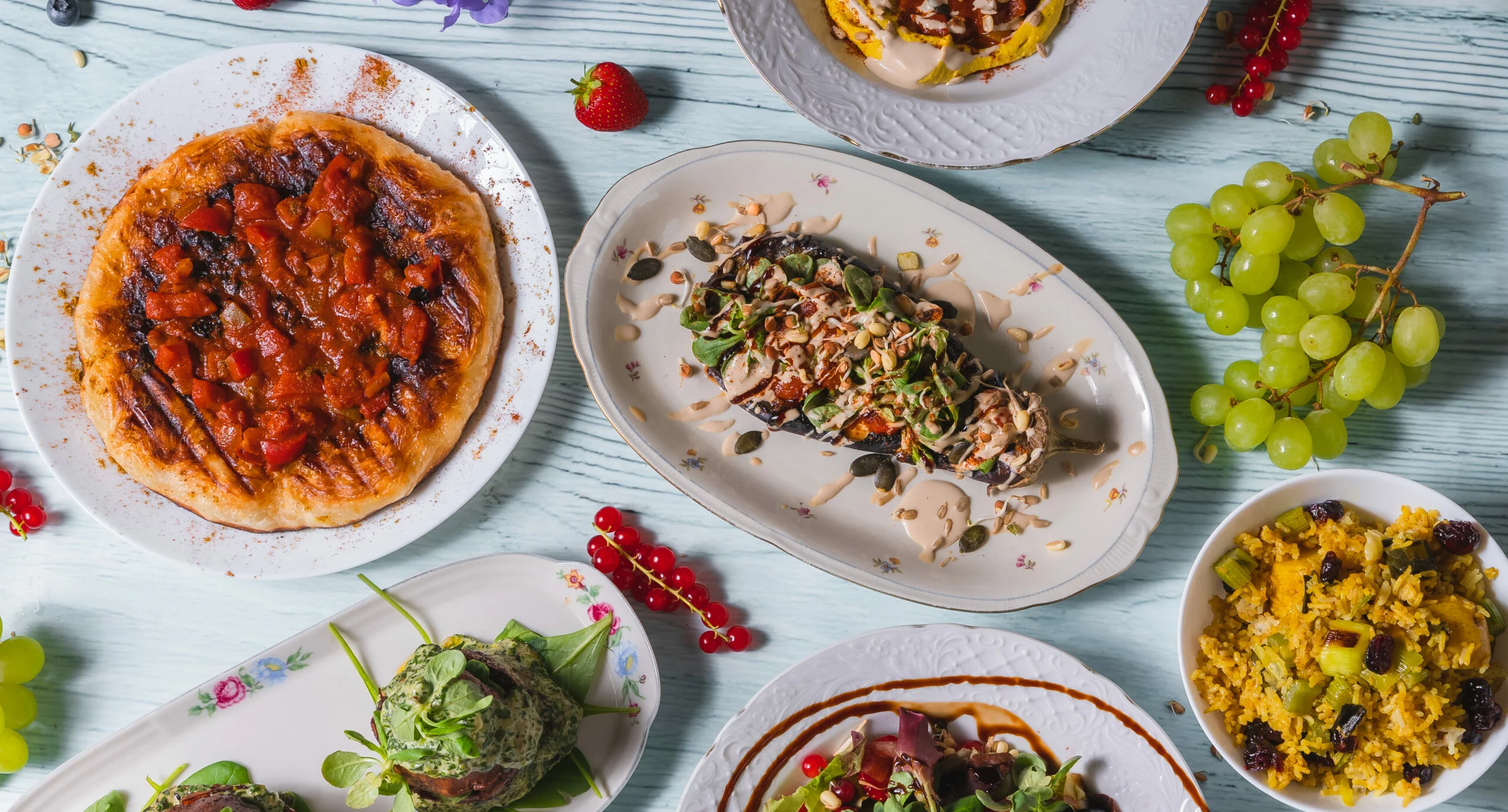 Mehrere Teller mit verschiedenen Gerichten, darunter ein Fladenbrot mit Tomatensauce, ein länglicher Teller mit Salat und Nüssen, Trauben und rote Johannisbeeren auf einem hellen Holztisch.
