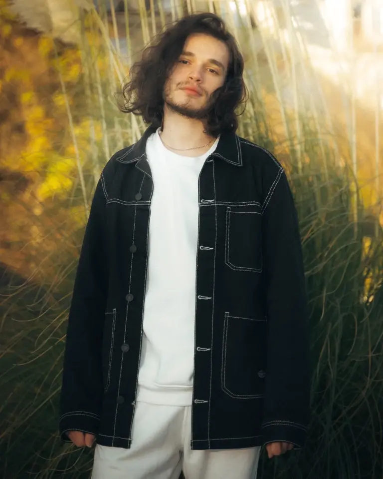 Person mit schulterlangen lockigen Haaren trägt schwarze Jacke mit weißen Nähten über weißem Oberteil und weißer Hose vor hohem Gras