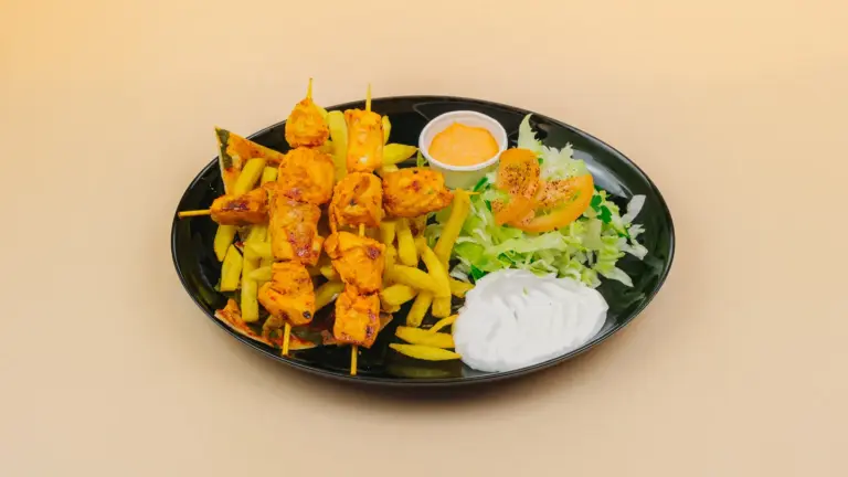 Schwarzer Teller mit Hähnchenspießen auf Pommes, Salat mit Tomatenscheibe und zwei Dips