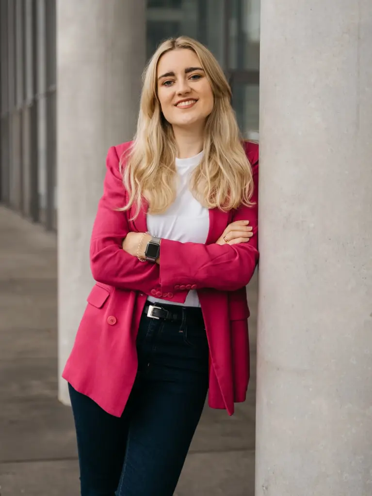 Frau mit langen blonden Haaren lehnt mit verschränkten Armen an einer Säule und trägt ein rotes Sakko, weißes T-Shirt und dunkle Hose
