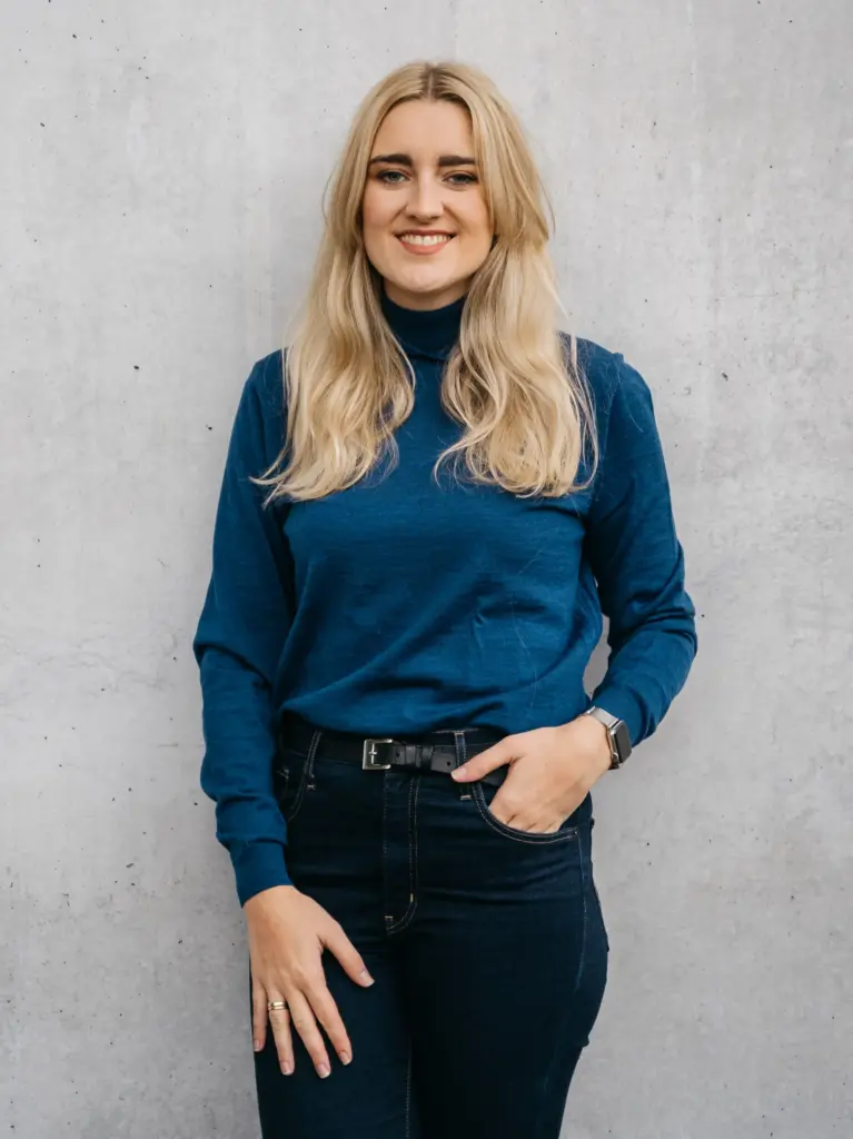 Frau mit langen blonden Haaren trägt einen dunkelblauen Rollkragenpullover und dunkle Jeans mit Gürtel vor einer hellen Betonwand