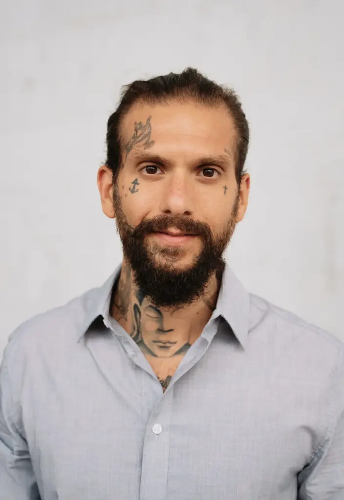 Oberkörper eines Mannes mit zusammengebundenem Haar, Hemd und sichtbaren Hals- und Brusttattoos vor hellem Hintergrund