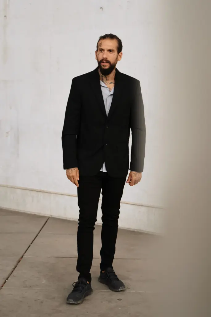 Ganzkörperaufnahme einer stehenden Person in schwarzem Anzug, schwarzer Hose und schwarzen Sportschuhen vor einer hellen Wand