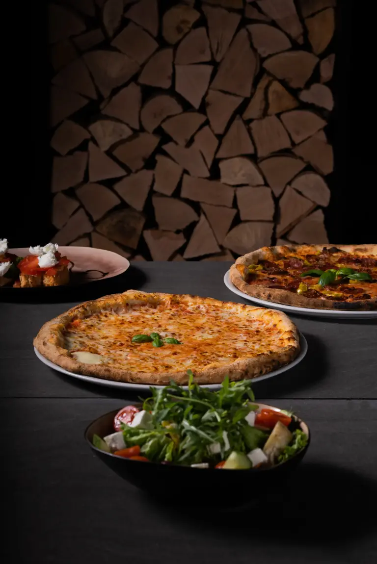 Zwei Pizzen auf Tellern, ein gemischter Salat in einer Schüssel und kleine Bruschetta vor einem Stapel Holzscheite