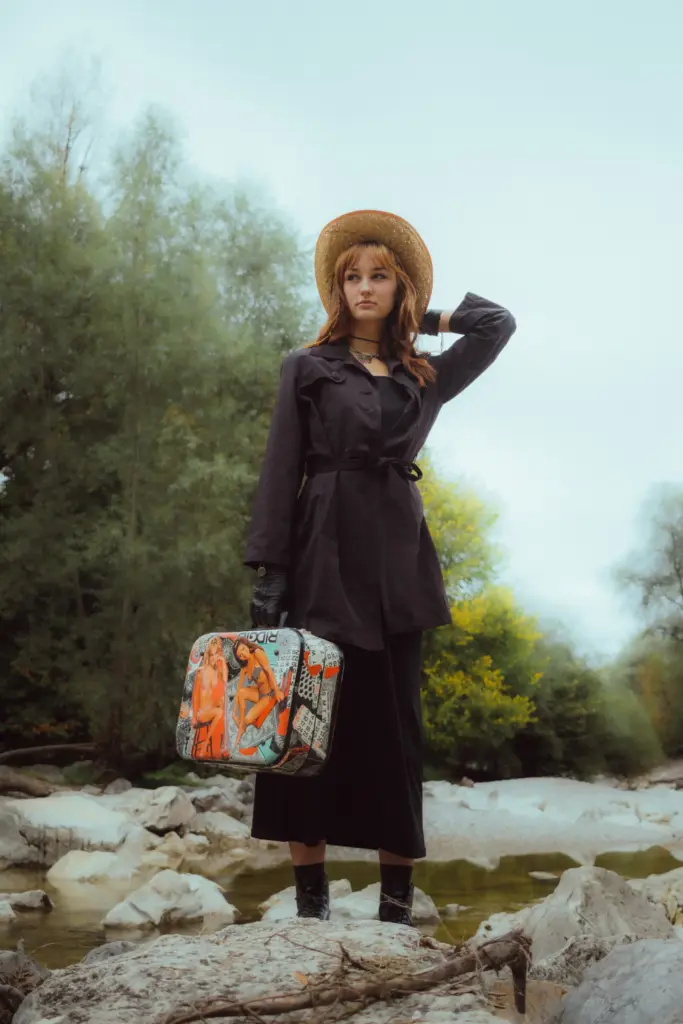 Frau mit Strohhut, schwarzem Mantel und Handschuhen steht auf Felsen an einem Fluss und hält einen Koffer mit buntem Motiv