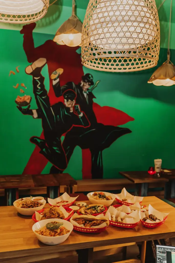 Tisch mit mehreren Schalen und Körben mit asiatischem Essen vor einer grünen Wand mit rotem Schatten und zwei Figuren in schwarzen Outfits und Hüten