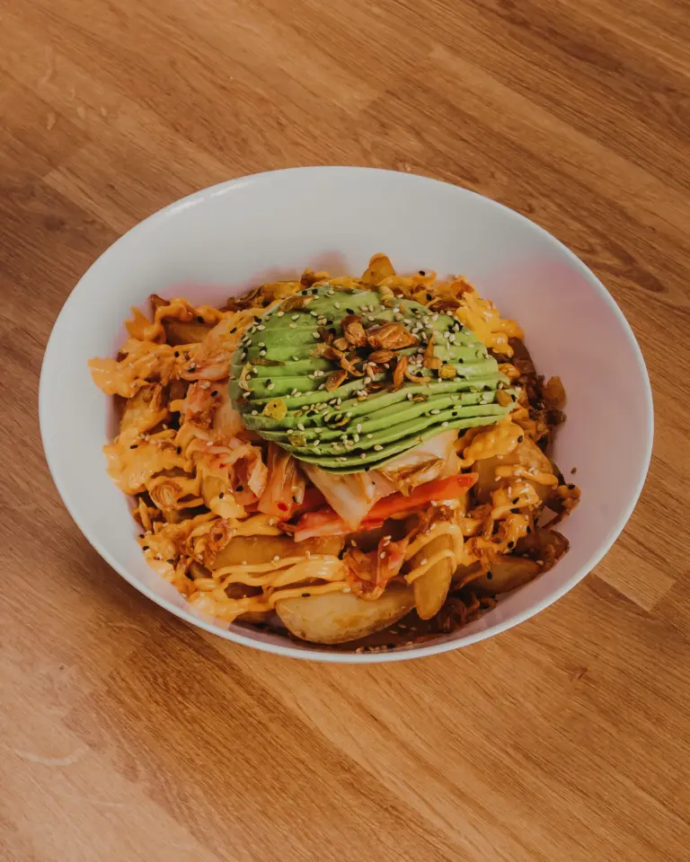Schale mit gewürzten Kartoffelspalten, Avocadoscheiben, Sesam und knusprigen Zwiebeln auf Holztisch