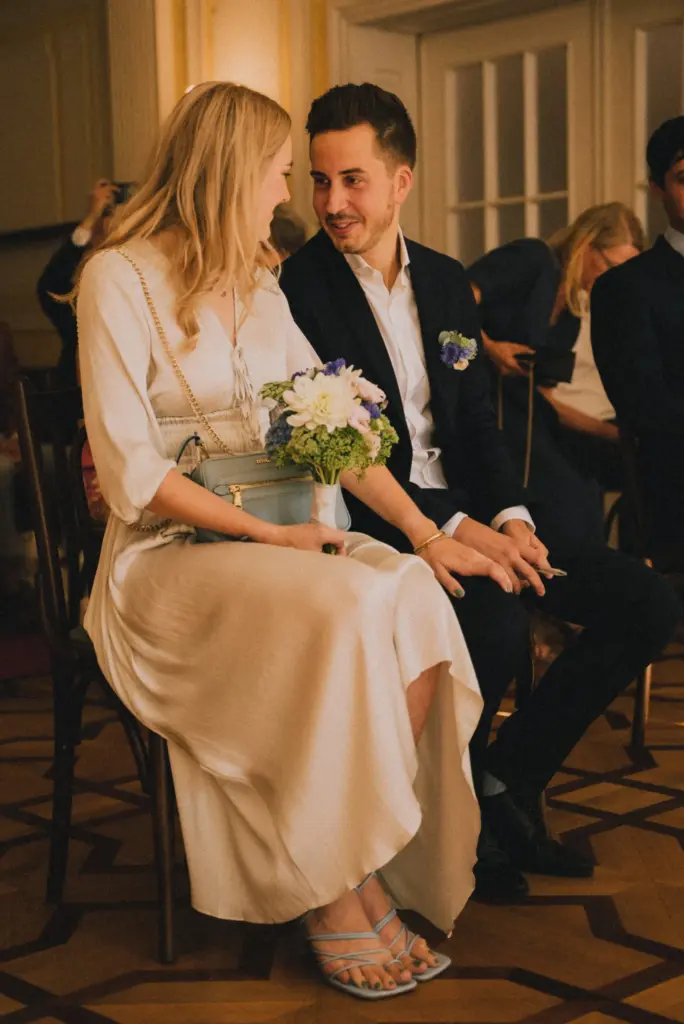 Paar sitzt nebeneinander auf Stühlen, die Frau trägt ein langes Kleid und hält einen Blumenstrauß, der Mann trägt einen Anzug mit Blumenanstecker