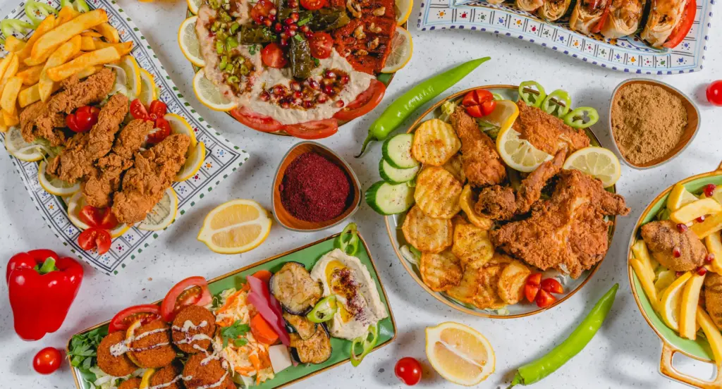 Verschiedene Teller mit frittiertem Hühnchen, Pommes, gegrilltem Gemüse, Salat und Gewürzen auf hellem Untergrund.