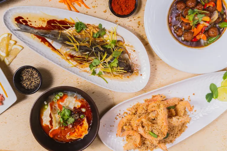Verschiedene asiatische Gerichte auf Tellern, darunter ein ganzer Fisch mit Kräutern, frittierte Garnelen mit Panade, eine Schüssel mit Teigtaschen in roter Sauce und eine Schüssel mit Fleischbällchen und Gemüse