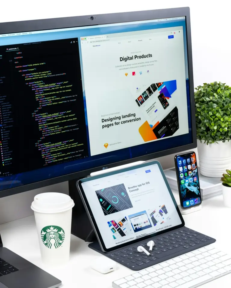 Arbeitsplatz mit großem Monitor, der links Programmiercode und rechts eine Webseite zu digitalen Produkten zeigt, davor Tablet mit Tastatur und App-Design, daneben Smartphone, zwei Pflanzen und ein Starbucks-Pappbecher